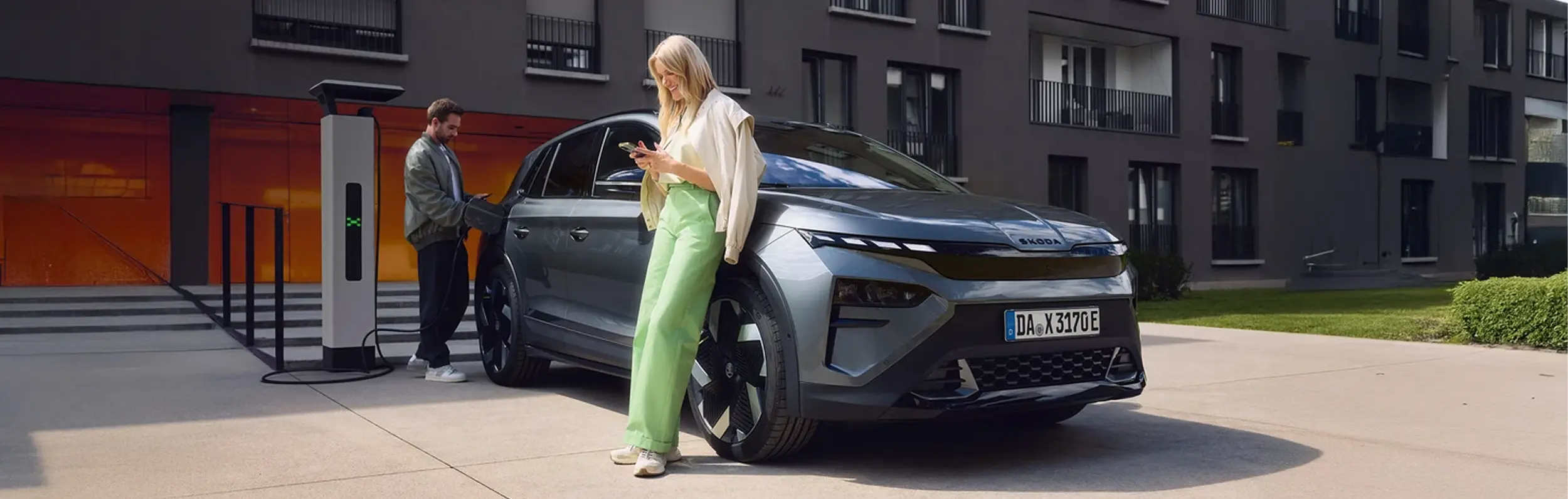 zu sehen ist ein blauer Skoda Enyaq von der Seite. Eine Frau lehnt sich an, ein Mann lädt das Auto.